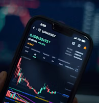 A close-up view of a smartphone displaying a cryptocurrency trading app with a graph showing the price movement of LUNA/USDT. The phone screen shows the current price, percentage change, and volume. The background is blurred with colorful digital charts, suggesting a trading environment.